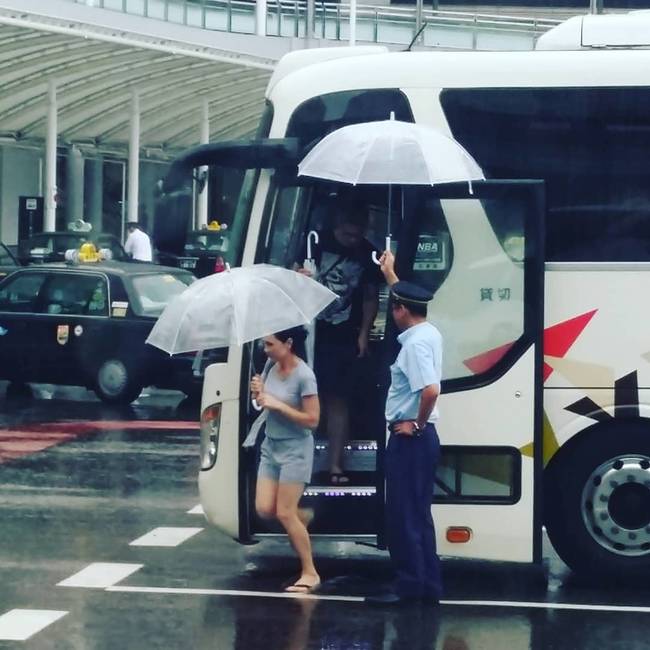 Egy japán buszvezető esernyőt tart az utasok feje fölé, hogy ne ázzanak meg az esőben.