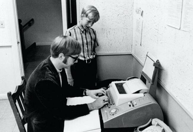Bill Gates csíkos ingben nézi, ahogy egy számítógépen dolgoznak. A fotó több mint 50 évvel ezelőtt készült.